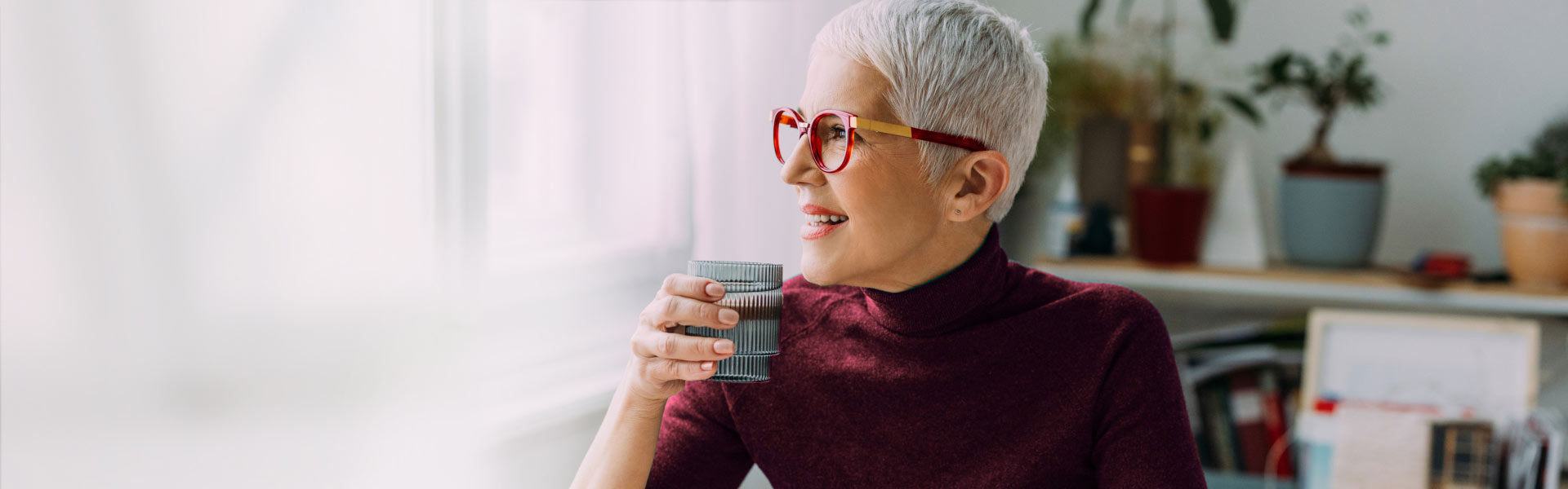 An older woman sitting, looking out a window, and drinking water.