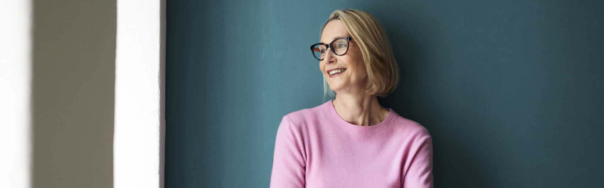 An older female leaning against a wall, looking into the distance and smiling.