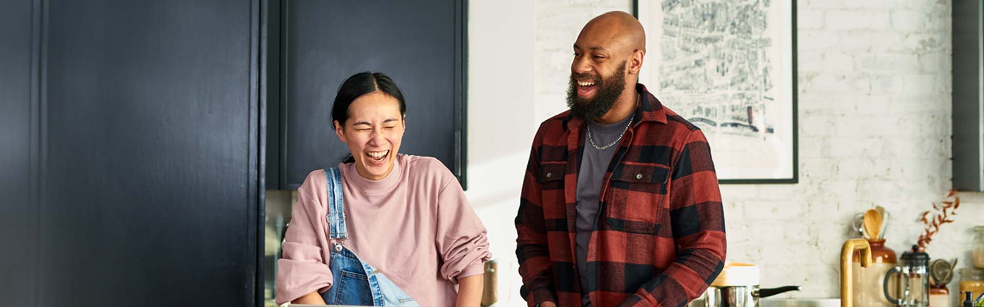 Two friends stand next to each other, laughing joyfully. The person on the left is wearing a light pink sweatshirt and the person on the right is wearing a black and red flannel shirt.